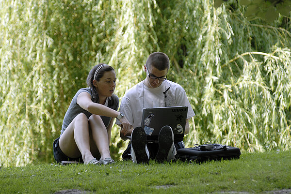 Foto: Zwei Personen sitzen auf einer Wiese im Freien. Sie arbeiten gemeinsam an einem Laptop, den eine der Personen auf dem Schoß hat.