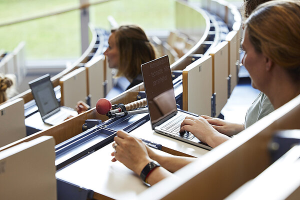 Foto: Personen in einem Hörsaal, die teilweise an Laptops arbeiten. Im Vordergrund ist eine Person zu sehen, die tippt und deren Bildschirm teilweise lesbar ist. Ein Langstock liegt auf einem der Tische.