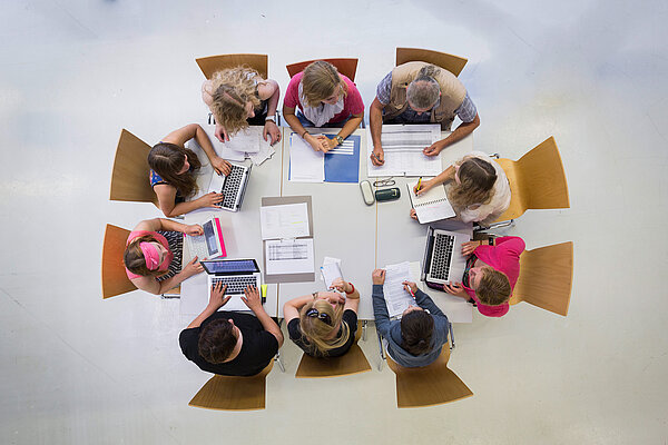 Foto, aufgenommen von oben: Gruppe von Menschen, die an einem großen Tisch sitzen. Auf dem Tisch liegen Laptops, Dokumente, Stifte und Notizbücher. Die Personen arbeiten, tippen auf Laptops, schreiben Notizen oder lesen Dokumente.