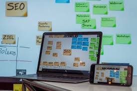 Foto: aufgeklappter Laptop, rechts daneben aufgestelltes Smartphone, beide Bildschirme bilden den Inhalt (Post-its in den Farben organe, blau und grün) des digitalen Whiteboards im Hintergrund ab